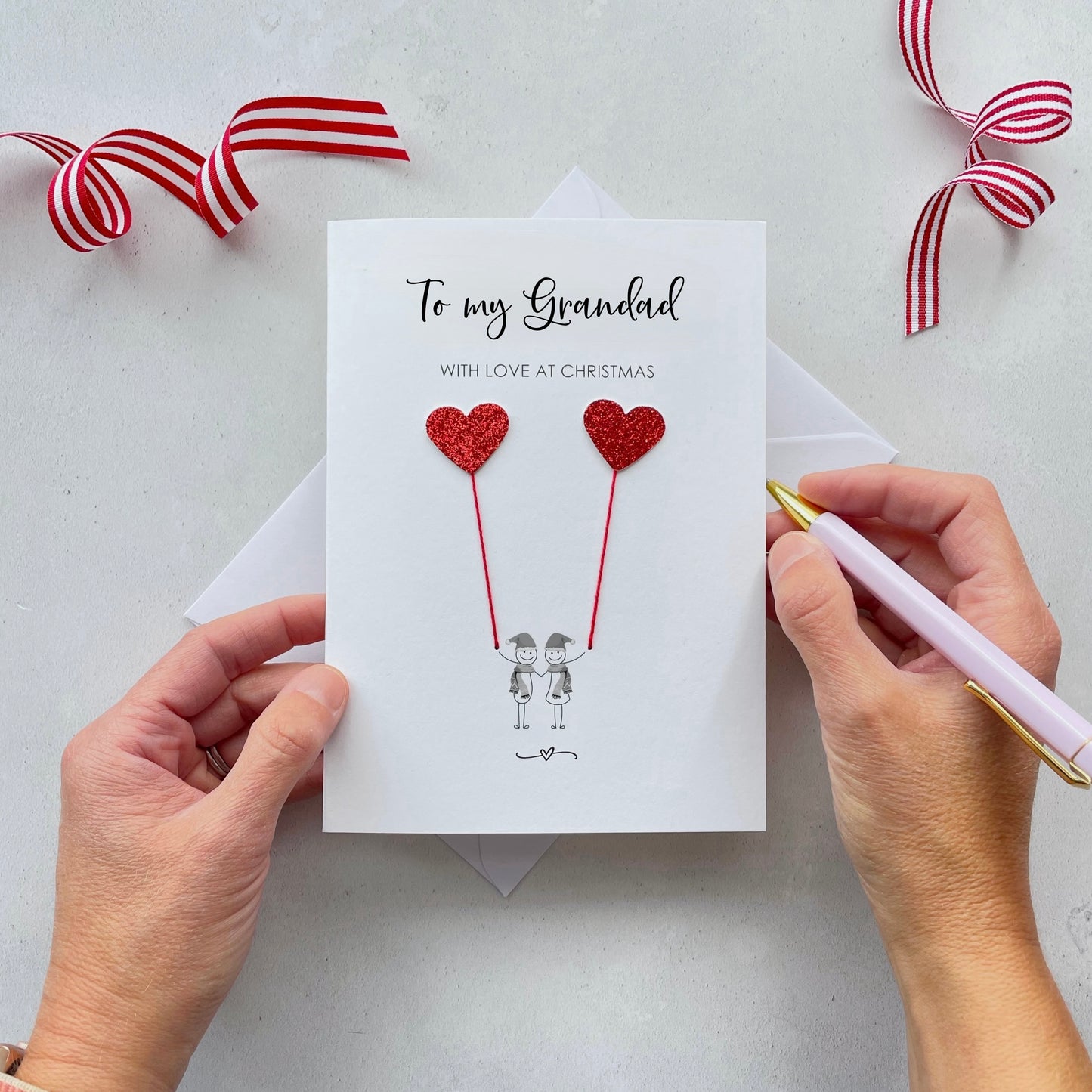 A Christmas card featuring a minimalist design with two figures wearing santa hats holding red glitter heart balloons. The text at the top reads: "To my Grandad, With Love at Christmas." The card is placed on a white background with a white envelope partially visible behind it. A hand is seen holding the card, while another hand holds a pink pen. A red and white striped ribbon is positioned in the top corners of the image for decoration.
