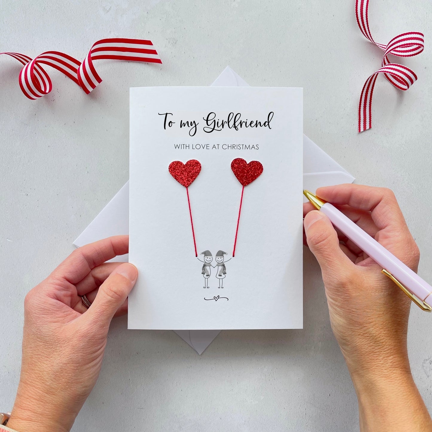 A Christmas card featuring a minimalist design with two figures wearing santa hats holding red glitter heart balloons. The text at the top reads: ‘To my Girlfriend, with love at Christmas’. The card is placed on a white background with a white envelope partially visible behind it. A hand is seen holding the card, while another hand holds a pink pen. A red and white striped ribbon is positioned in the top corners of the image for decoration.
