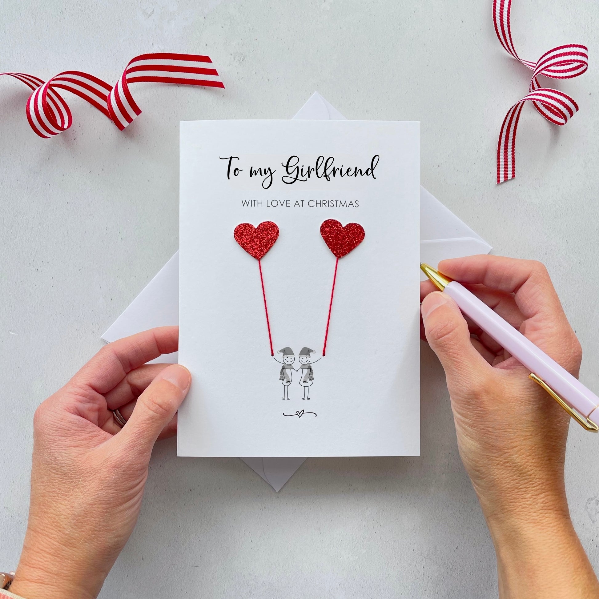 A Christmas card featuring a minimalist design with two figures wearing santa hats holding red glitter heart balloons. The text at the top reads: ‘To my Girlfriend, with love at Christmas’. The card is placed on a white background with a white envelope partially visible behind it. A hand is seen holding the card, while another hand holds a pink pen. A red and white striped ribbon is positioned in the top corners of the image for decoration.