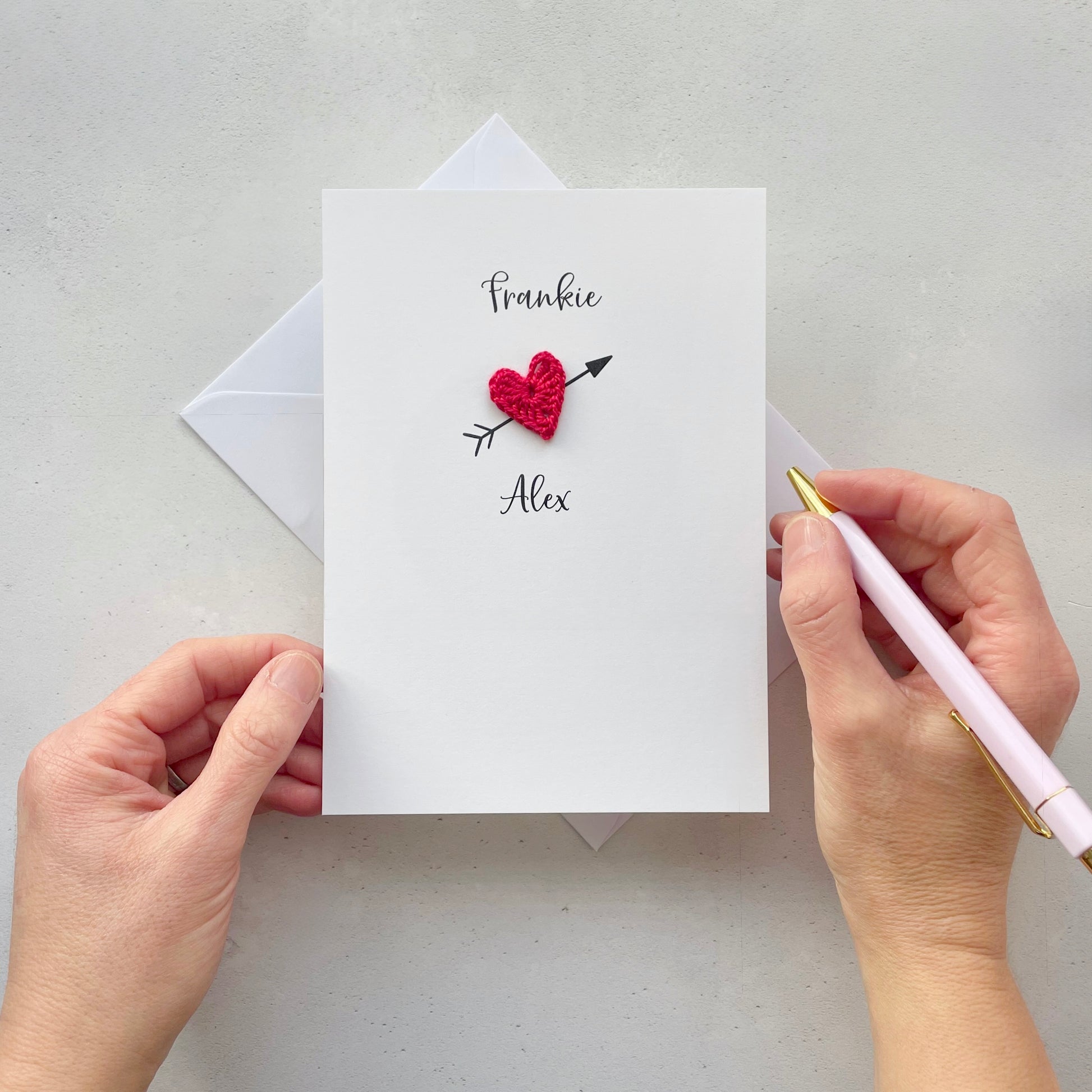 A white card with a red crochet heart in the centre. An arrow has been printed behind the heart so it looks like a cupid arrow. the name Frankie is printed above the heart and the name Alex is printed below the heart. these names have been printed in black cursive text. 
