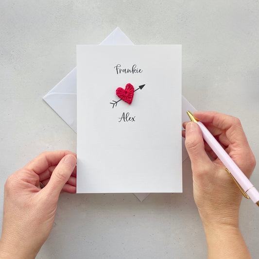 A white card with a red crochet heart in the centre. An arrow has been printed behind the heart so it looks like a cupid arrow. the name Frankie is printed above the heart and the name Alex is printed below the heart. these names have been printed in black cursive text. 