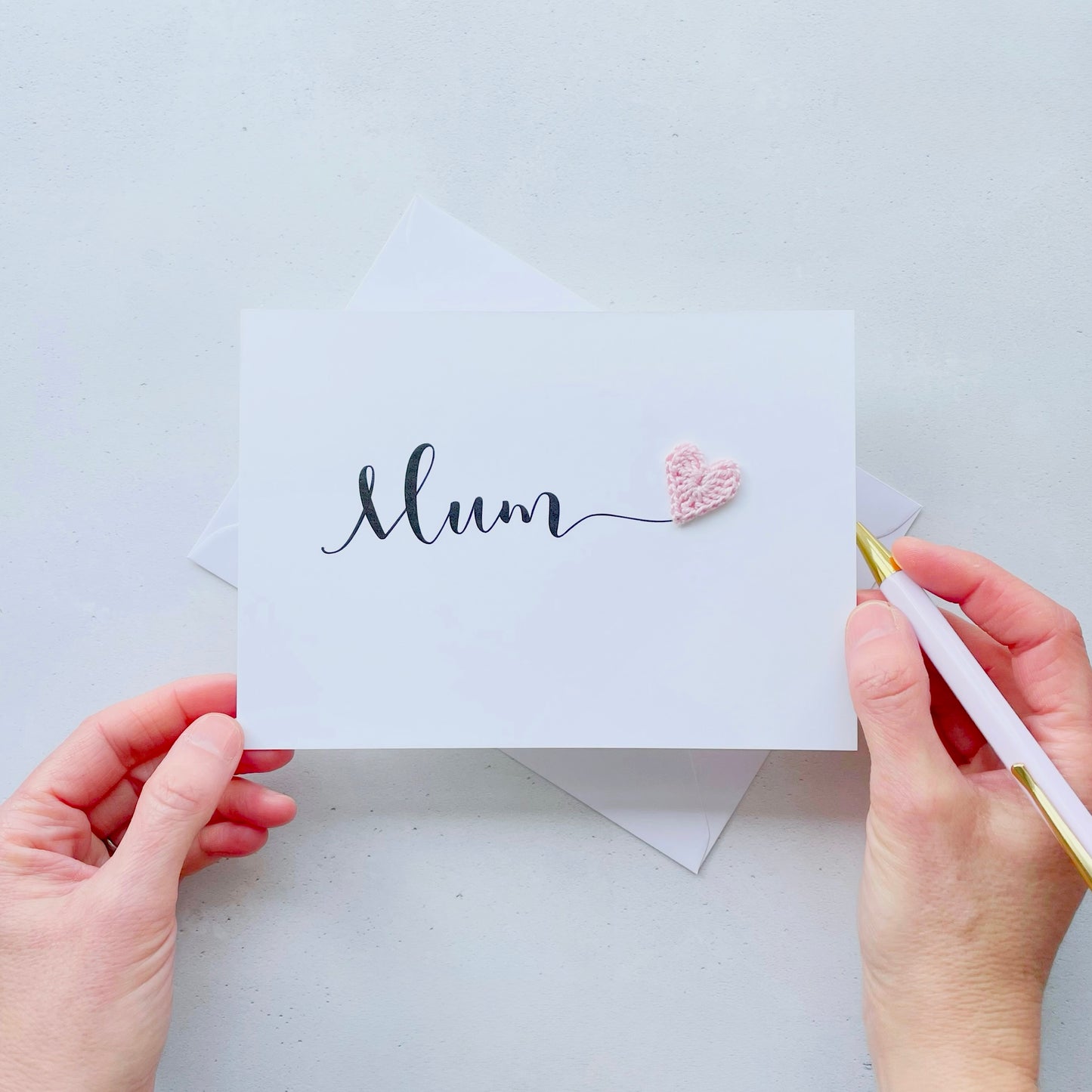 A white card with the word 'Mum' printed across the middle in black script text. There is a bright pink crochet heart stuck onto the end of the word 'Mum'
