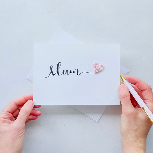 A white card with the word 'Mum' printed across the middle in black script text. There is a bright pink crochet heart stuck onto the end of the word 'Mum'
