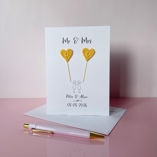 The image shows a beautiful personalised wedding card with little yellow crochet hearts, these are attached by cotton yarn, resembling balloons being held by a hand-drawn couple underneath. The text on the card reads 'Mr & Mrs' at the top, followed by the couples names and wedding date at the bottom.