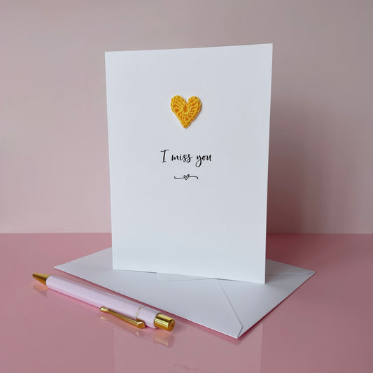 Greeting card with a yellow heart and 'I miss you' text on a pink surface with an envelope and pen.