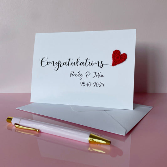 A white card with 'Congratulations' printed across the card in large black script text. There is a red crochet heart at the end of that word and underneath 'Congratulations', you've got the two names and their wedding date. 