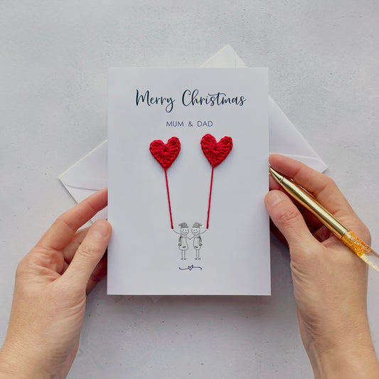 A white Christmas card featuring a line drawn couple holding hands and each holding a red crochet heart balloon. ‘Merry Christmas’ is printed at the top of the card in black cursive text and under that ‘Mum & Dad’ is printed in smaller text. The couple are drawn wearing winter scarves and santa hats.