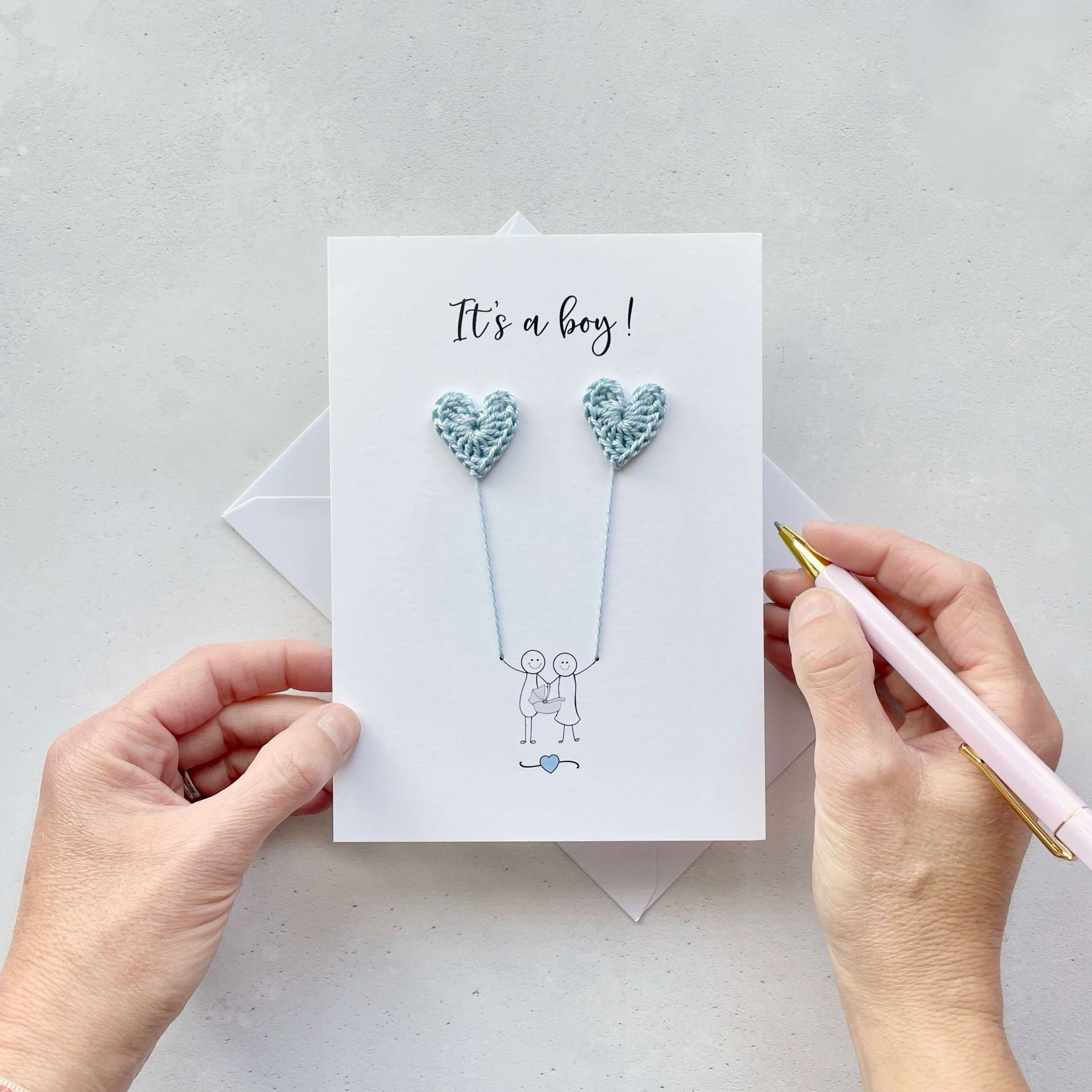 It's a boy is printed at the top of the card in black cursive text. A line drawn illustration of a couple appears on the card - they are holding a baby car seat. The are each holding a pale blue crochet heart balloon. 