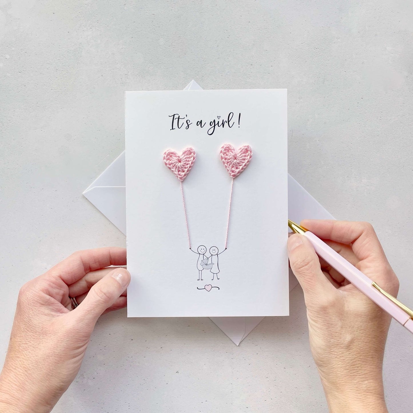 It's a girl is printed at the top of the card in black cursive text. A line drawn illustration of a couple appears on the card - they are holding a baby car seat. The are each holding a pale pink crochet heart balloon. 