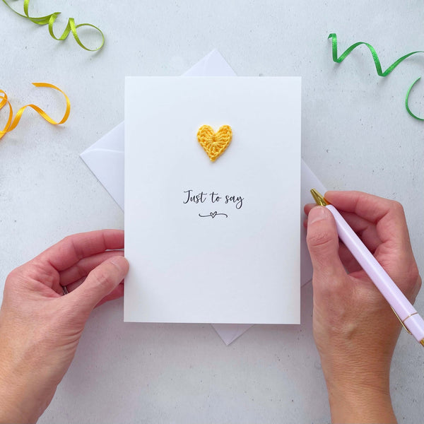 A white portrait-style card featuring the words ‘Just to say’ in elegant black cursive at the center. Below the text, a small black squiggle and heart design adds a playful touch. Above the words, a handmade yellow crochet heart is carefully attached, providing a pop of colour and texture.