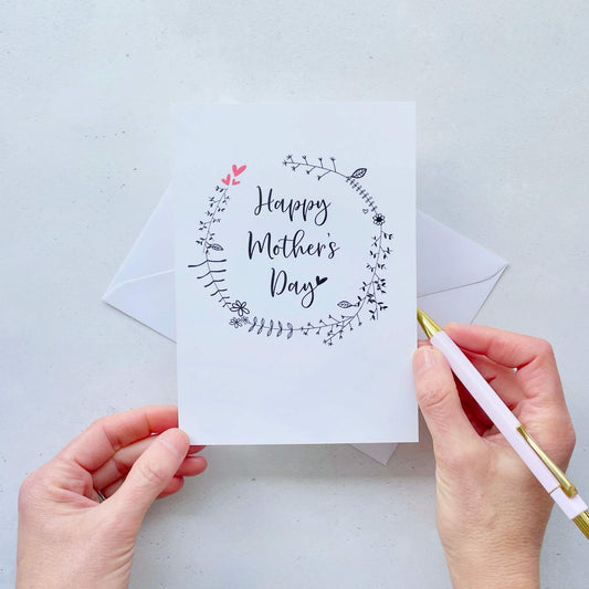 A white Mother’s Day card. Black line drawn foliage is printed on teh card in a circle pattern. The words ‘Happy Mother’s Day’ has been printed inside the circle in black cursive text. 