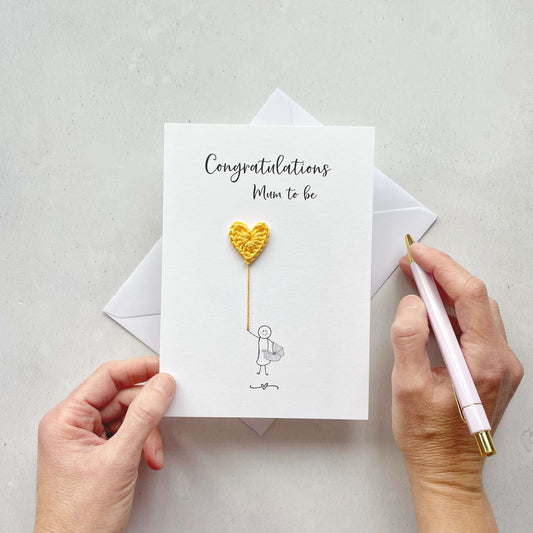 A white card with 'Congratulations Mum to be' printed at the top. An illustration of a Mum holding a bady carrier is holding a yellow crochet heart balloon. 
