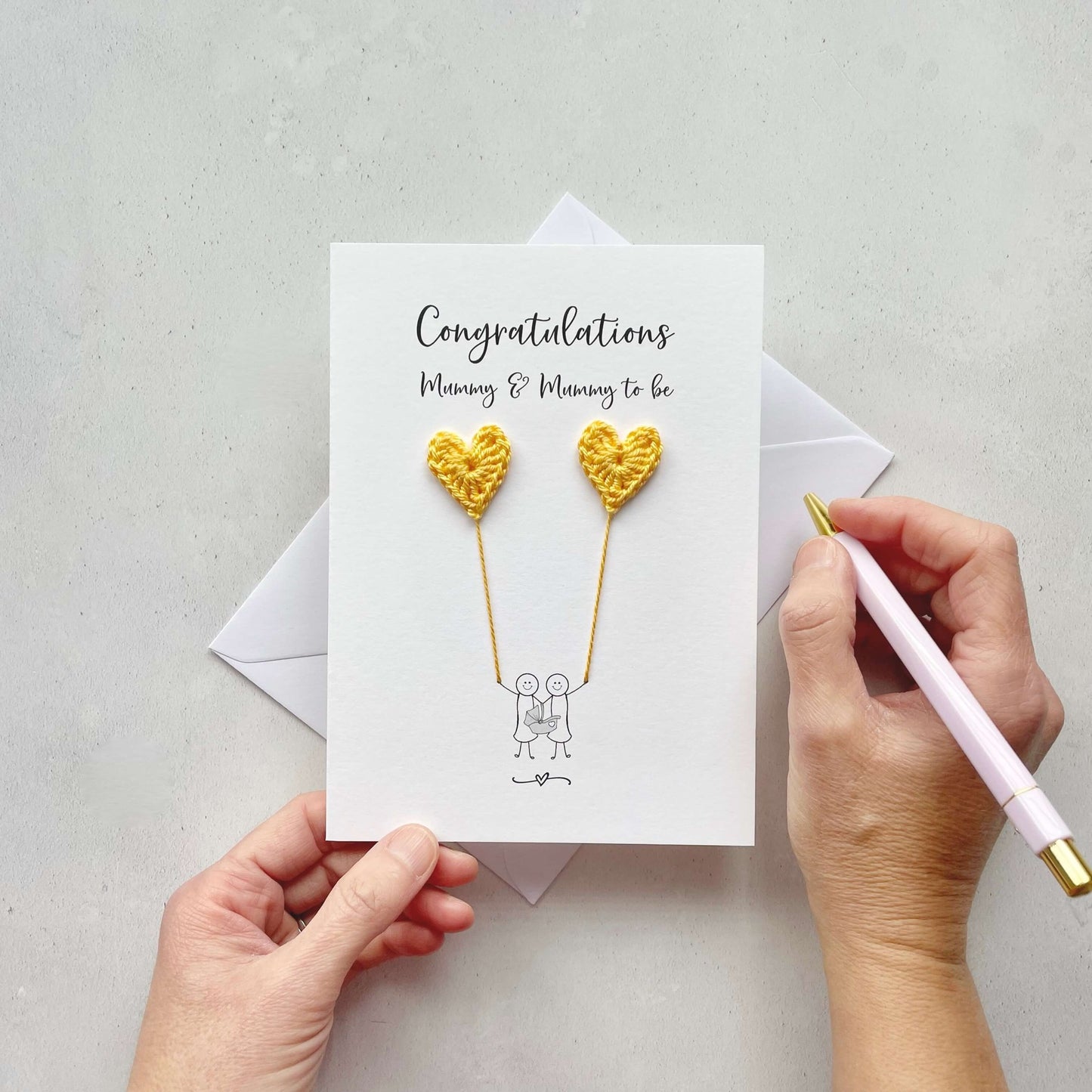 A white card with 'Congratulations Mummy & mummy to be' printed at the top. the illustrated couple are each holding a yellow crochet heart, this looks like they are holding balloons. 