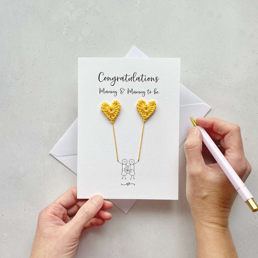 A white card with 'Congratulations Mummy & mummy to be' printed at the top. the illustrated couple are each holding a yellow crochet heart, this looks like they are holding balloons. 