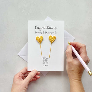 A white card with 'Congratulations Mummy & mummy to be' printed at the top. the illustrated couple are each holding a yellow crochet heart, this looks like they are holding balloons. 