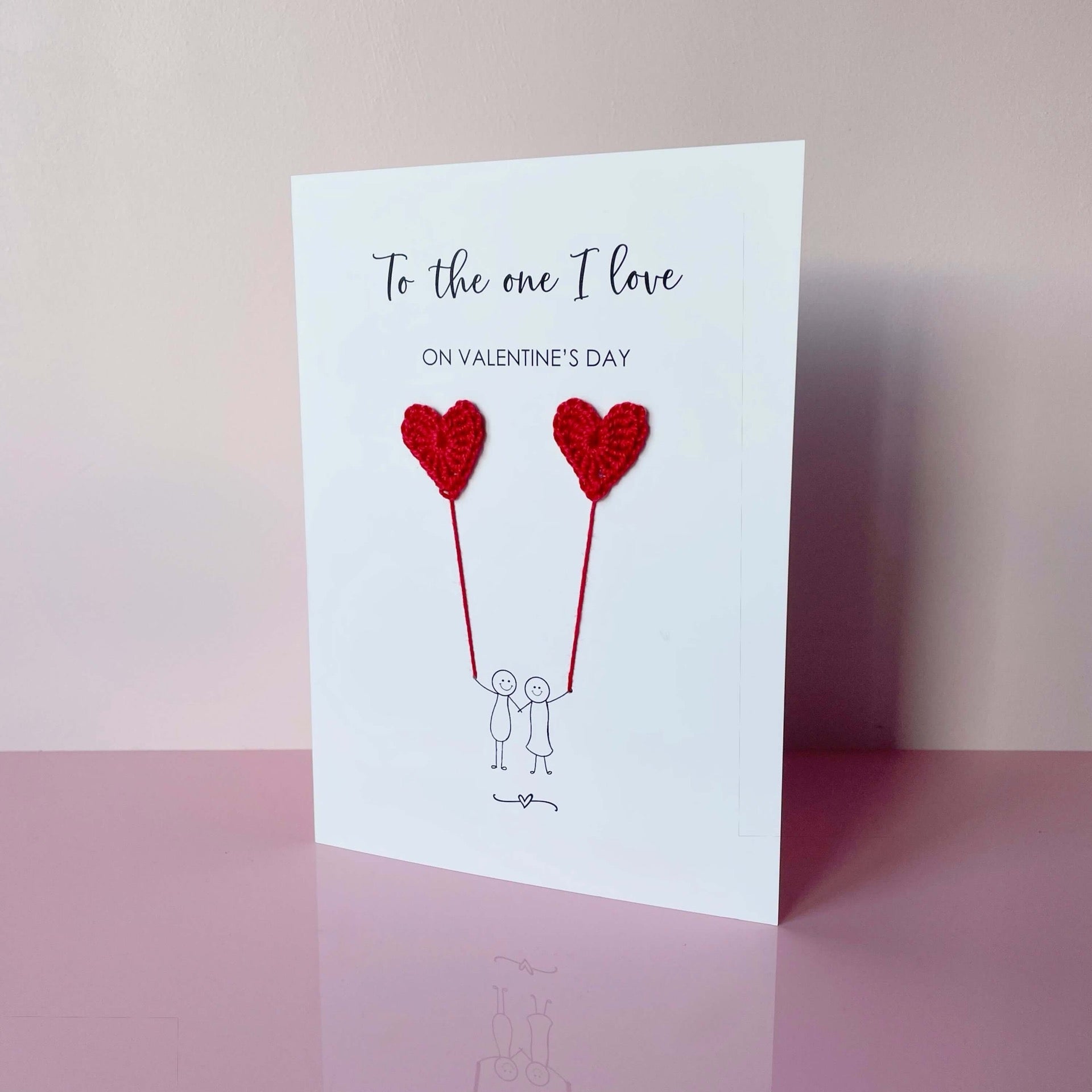 A Valentines card featuring a minimalist design with two figures holding red sparkly card heart shaped balloons. The text at the top reads: ‘To the one I love on Valentine’s day’. The card is photographed on a pale pink background.