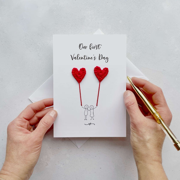 A white card featuring a line drawn couple holding hands and each holding a red crochet heart balloon.  ‘Our first Valentine’s Day’ is printed at the top of the card in black cursive text.