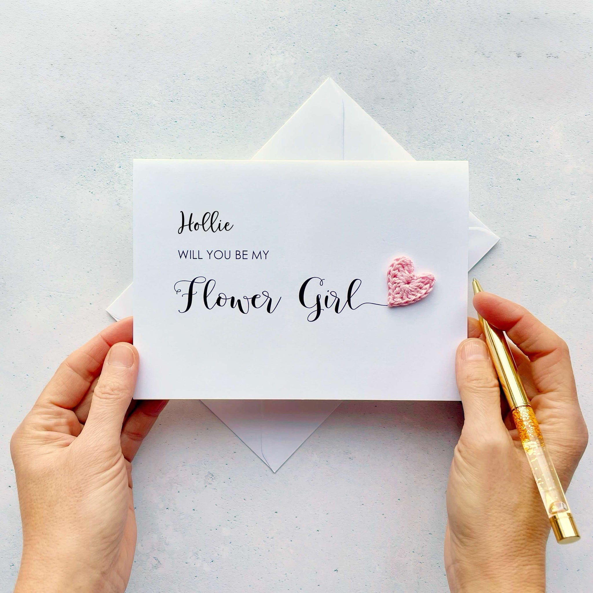 Will you be my Flower Girl has been printed on a white card (landscape). Flower girl has been printed larger than all other text on the card, it has a pale pink crochet heart at the end of the word. This design has been personalised with a name, the name appears at the top of the card in black cursive text.