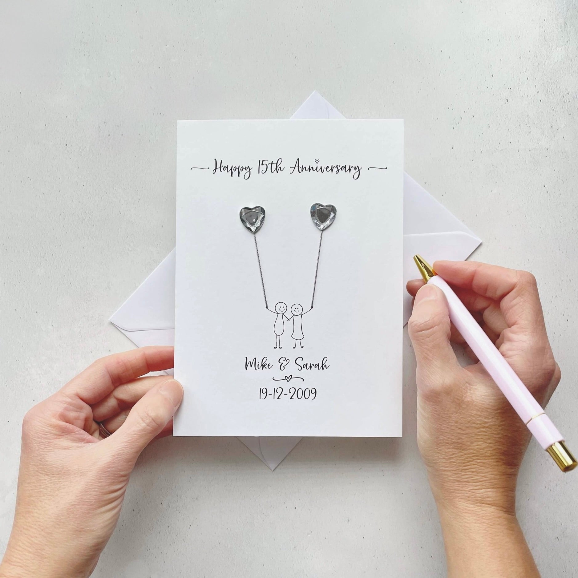 Handmade anniversary card embelished with 2 heart shaped plastic gems, both attached by cotton yarn, resembling balloons being held by a couple underneath. Happy 15th Anniversary is printed at the top, the couples names/ wedding date at the bottom.