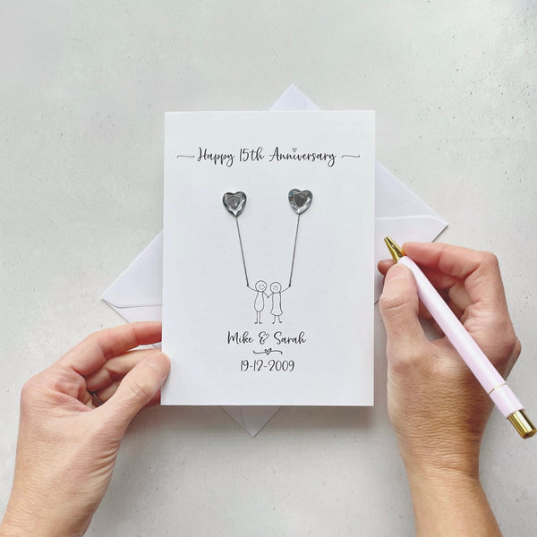 Handmade anniversary card embelished with 2 heart shaped plastic gems, both attached by cotton yarn, resembling balloons being held by a couple underneath. Happy 15th Anniversary is printed at the top, the couples names/ wedding date at the bottom.