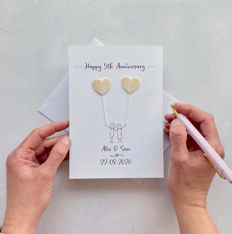 A white card featuring a line drawn couple holding hands and each holding a heart shaped balloon which is made from balsa wood. Happy 5th Anniversary is printed at the top of the card and the couples names and wedding date are printed at the bottom.