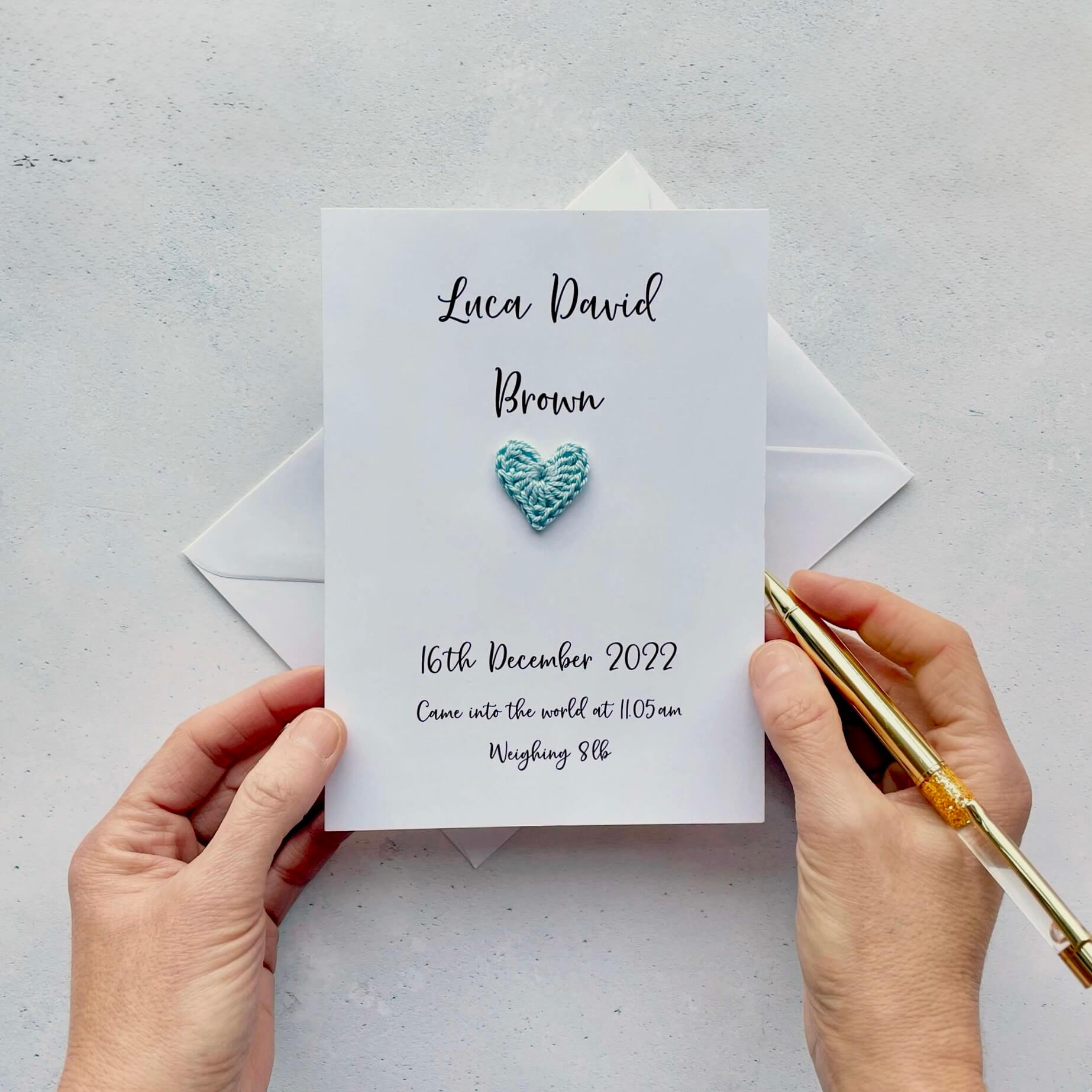 Newborn announcement card with blue heart design held by hands on a light gray background