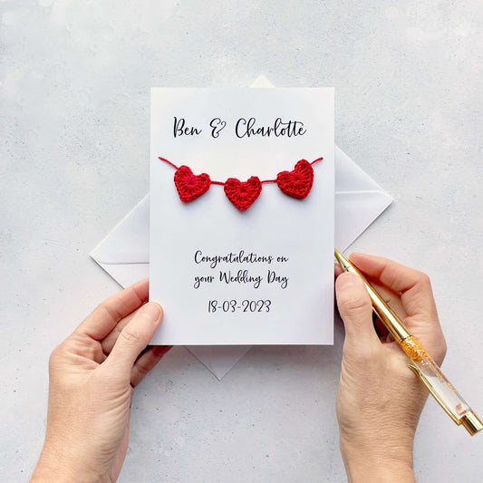 A white card with a string of 3 red crochet hearts across the front. Above the hearts are the two names and at the bottom of the card is 'Congratulations on your wedding day' and the wedding date. 