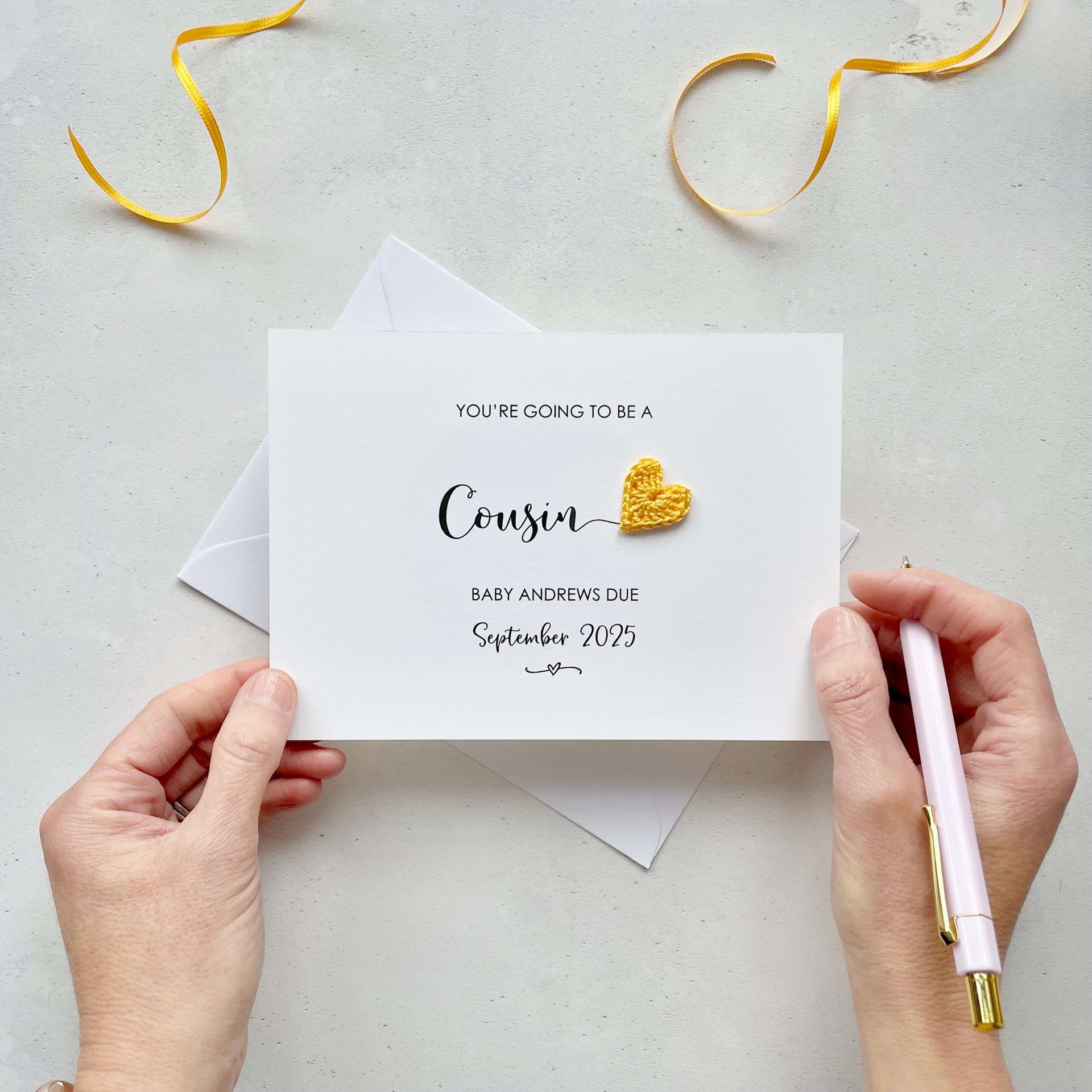 A landscape pregnancy announcement card with the text “You’re going to be a Cousin” in elegant black font. Below, it says “Baby Andrews due September 2025,” with a small decorative flourish. The card features a small crocheted yellow heart.