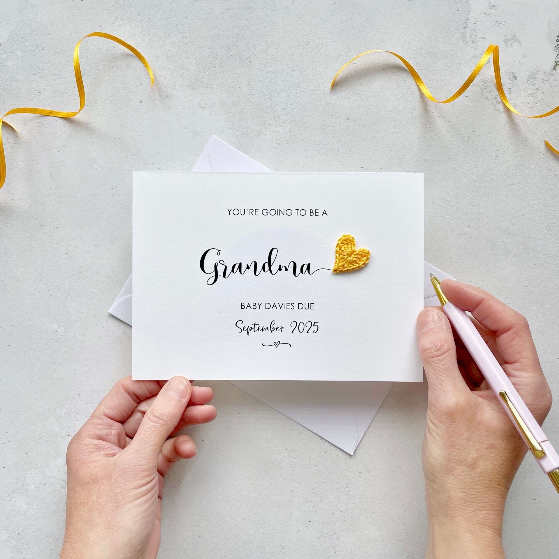 A landscape pregnancy announcement card with the text “You’re going to be a Grandma” in elegant black font. Below, it says “Baby Davies due September 2025,” with a small decorative flourish. The card features a small crocheted yellow heart.