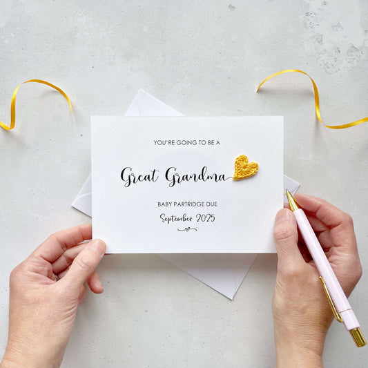 A landscape pregnancy announcement card with the text “You’re going to be a Great Grandma” in elegant black font. Below, it says “Baby Partridge due September 2025,” with a small decorative flourish. The card features a small crocheted yellow heart.