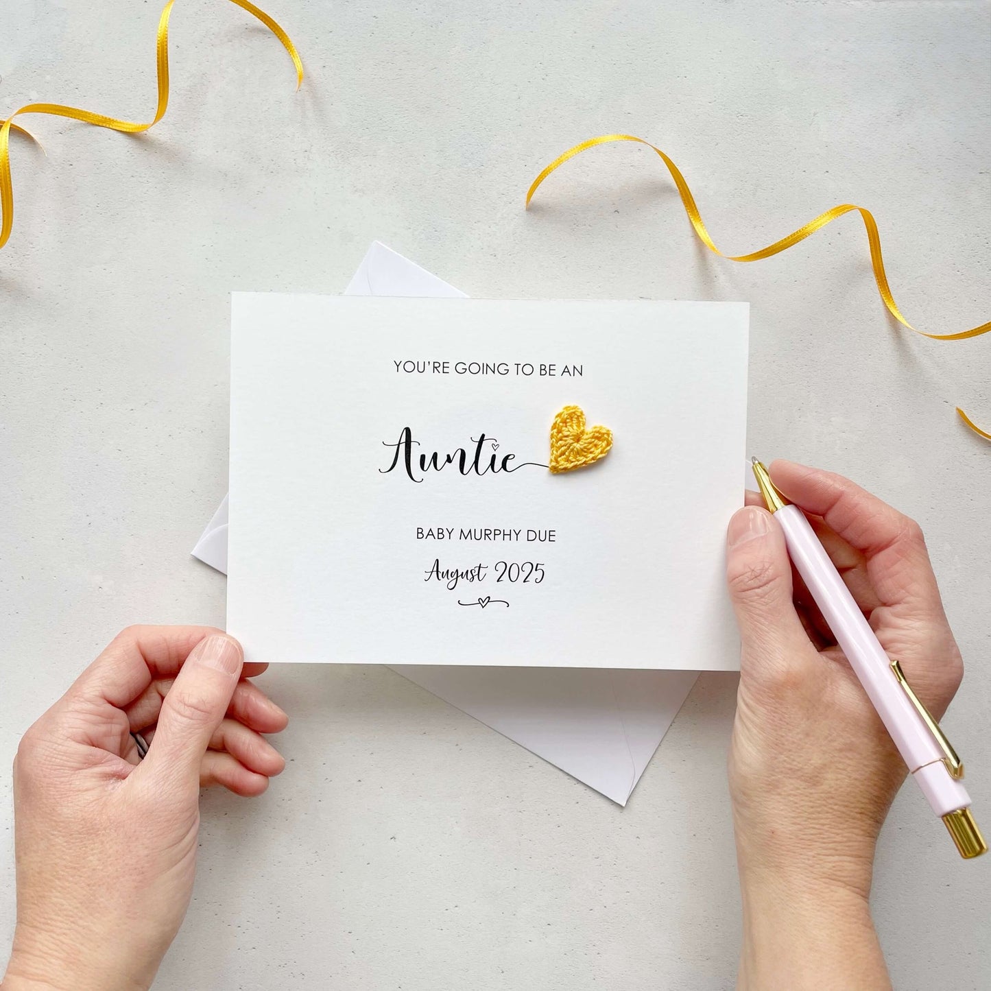 A landscape pregnancy announcement card with the text “You’re going to be an Auntie” in elegant black font. Below, it says “Baby Murphy due August 2025,” with a small decorative flourish. The card features a small crocheted yellow heart.