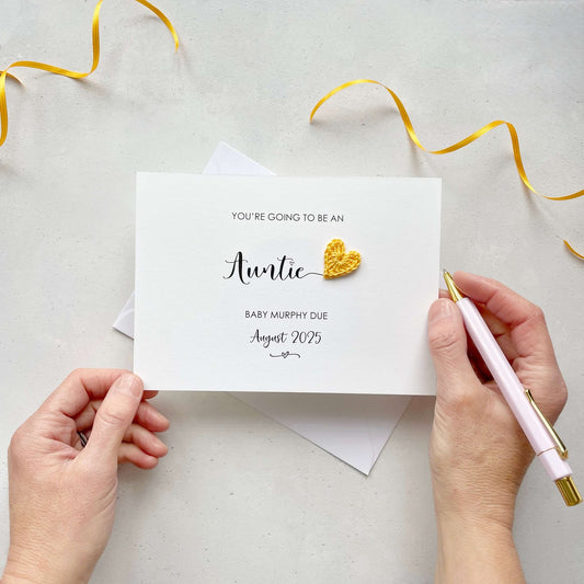 A landscape pregnancy announcement card with the text “You’re going to be an Auntie” in elegant black font. Below, it says “Baby Murphy due August 2025,” with a small decorative flourish. The card features a small crocheted yellow heart.