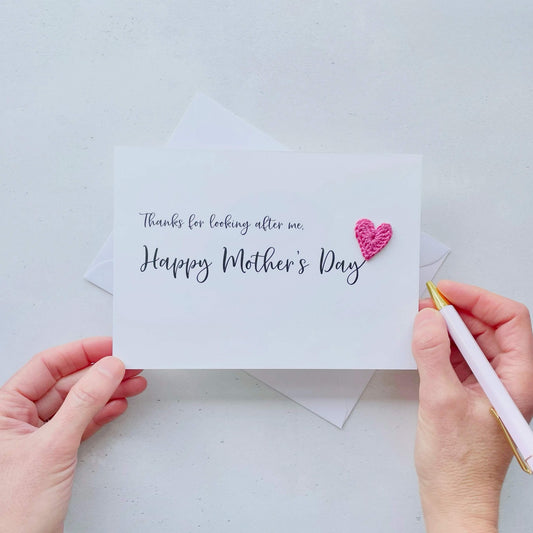 A white Mother’s Day card that has ‘Thanks for looking after me, Happy Mother’s Day’ printed on the card in black cursive text. A bright pink crochet heart has been attatched to the ‘y’ at the end. 