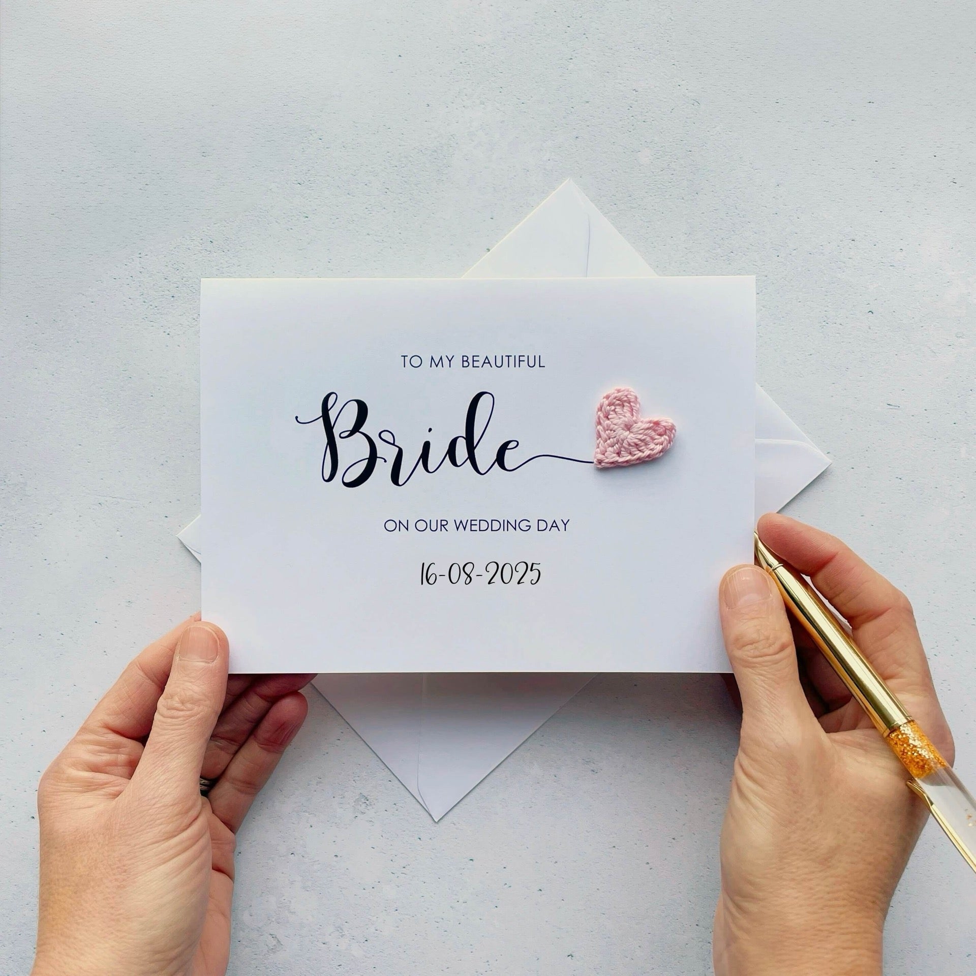 A white card with 'To my beautiful Bride on our wedding day' printed on the front of the card along with the wedding date. The word 'Bride' appears much larger than the rest and there is a pale pink crochet heart at the end of the letter 'e'