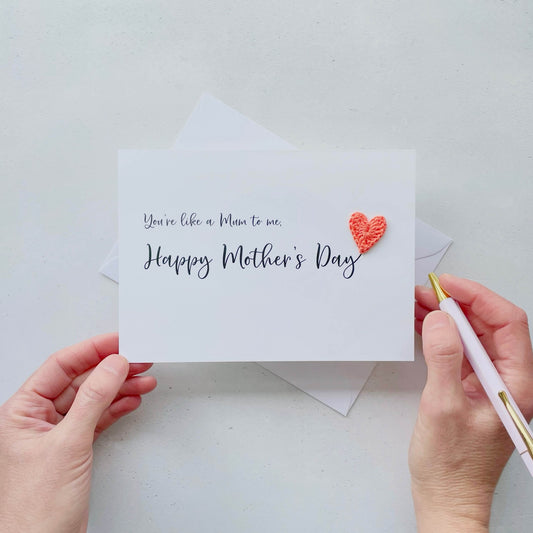 A white Mother’s Day card that has ‘You’re like a Mum to me , Happy Mother’s Day’ printed on the card in black cursive text. A bright pink crochet heart has been attatched to the ‘y’ at the end. 