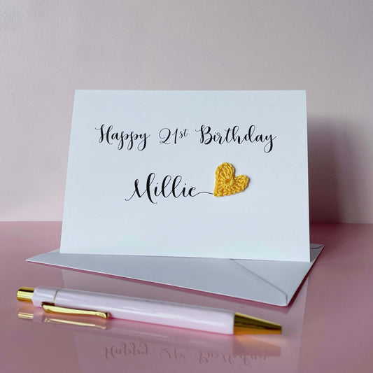 A white birthday card with 'Happy 21st Birthday Millie' printed on it in black cursive text. There is a yellow crochet heart at the end of the name Millie. 