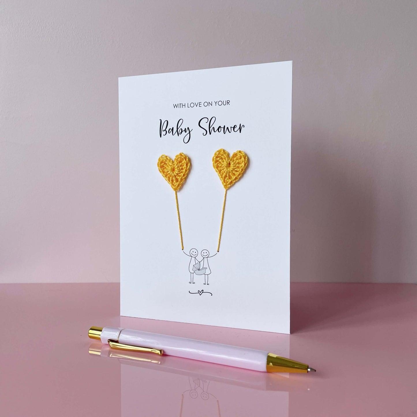 a white card with 'with love on your baby shower' printed at the top. A line drawn couple holding a baby carrier are printed at the bottom of the card - they are each holding a handmade yellow crochet heart balloon.