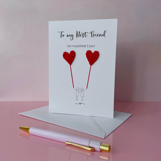 A Valentines card featuring a minimalist design with two figures holding red crochet heart balloons. The text at the top reads: ‘To my Best friend on Valentine’s day’. 