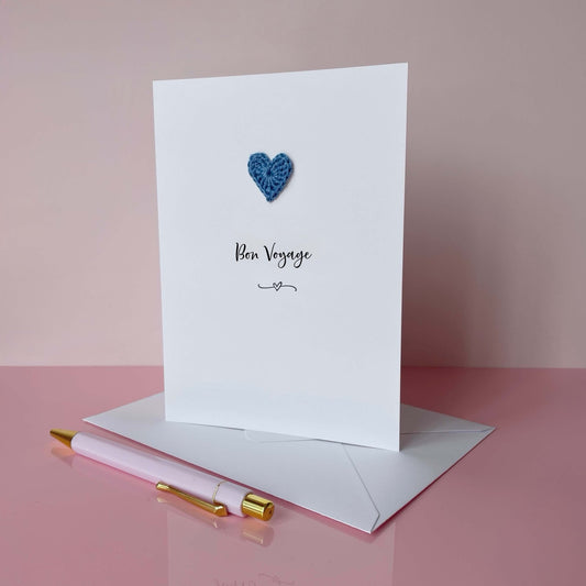 Greeting card with a blue crochet heart and text on a pink background, 'Bon Voyage' is printed on the card in lovely black cursive text. 