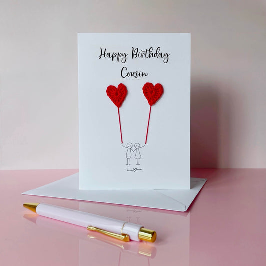 A white birthday card featuring a line drawn couple holding hands and each holding a red crochet heart balloon.  ‘Happy Birthday Cousin' is printed at the top of the card in black cursive text.