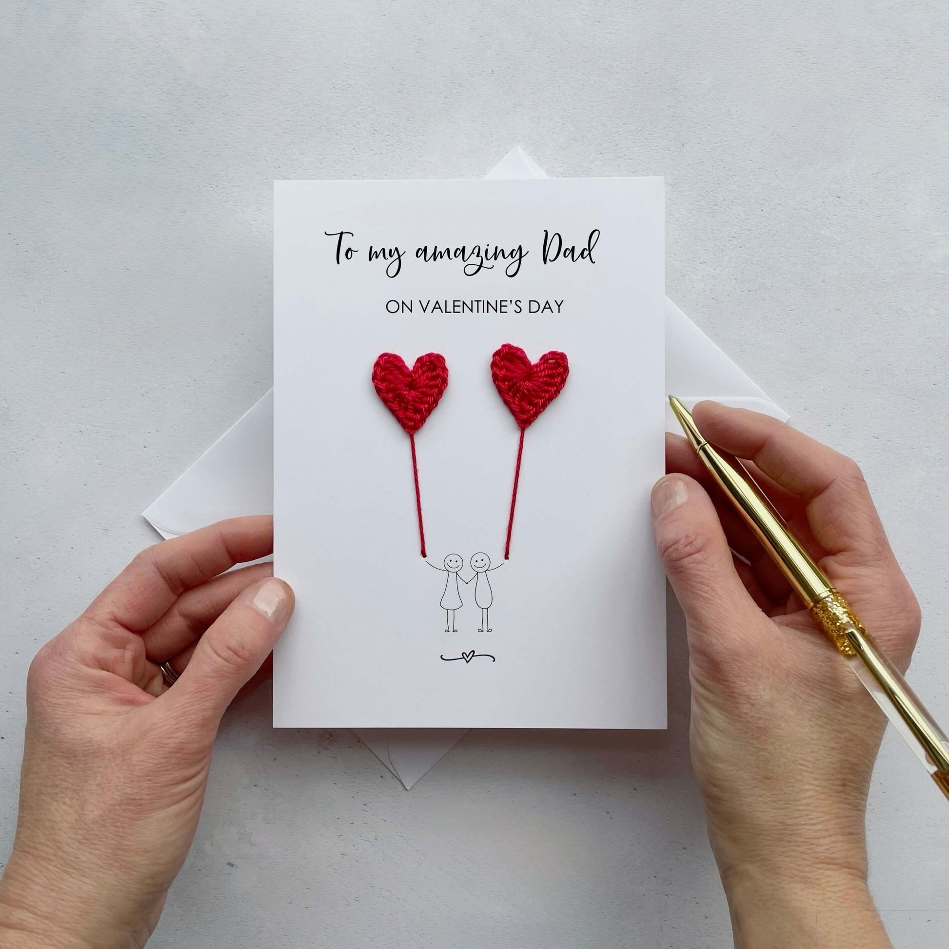 A white card featuring a line drawn couple holding hands and each holding a red crochet heart balloon.  ‘To my amazing Dad on Valentine’s Day’ is printed at the top of the card in black cursive text.