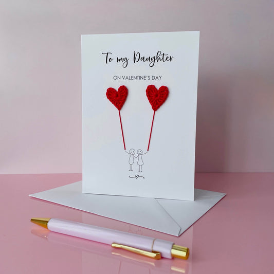 A white card featuring a line drawn couple holding hands and each holding a red crochet heart balloon.  ‘To my Daughter on Valentine’s Day’ is printed at the top of the card in black cursive text.