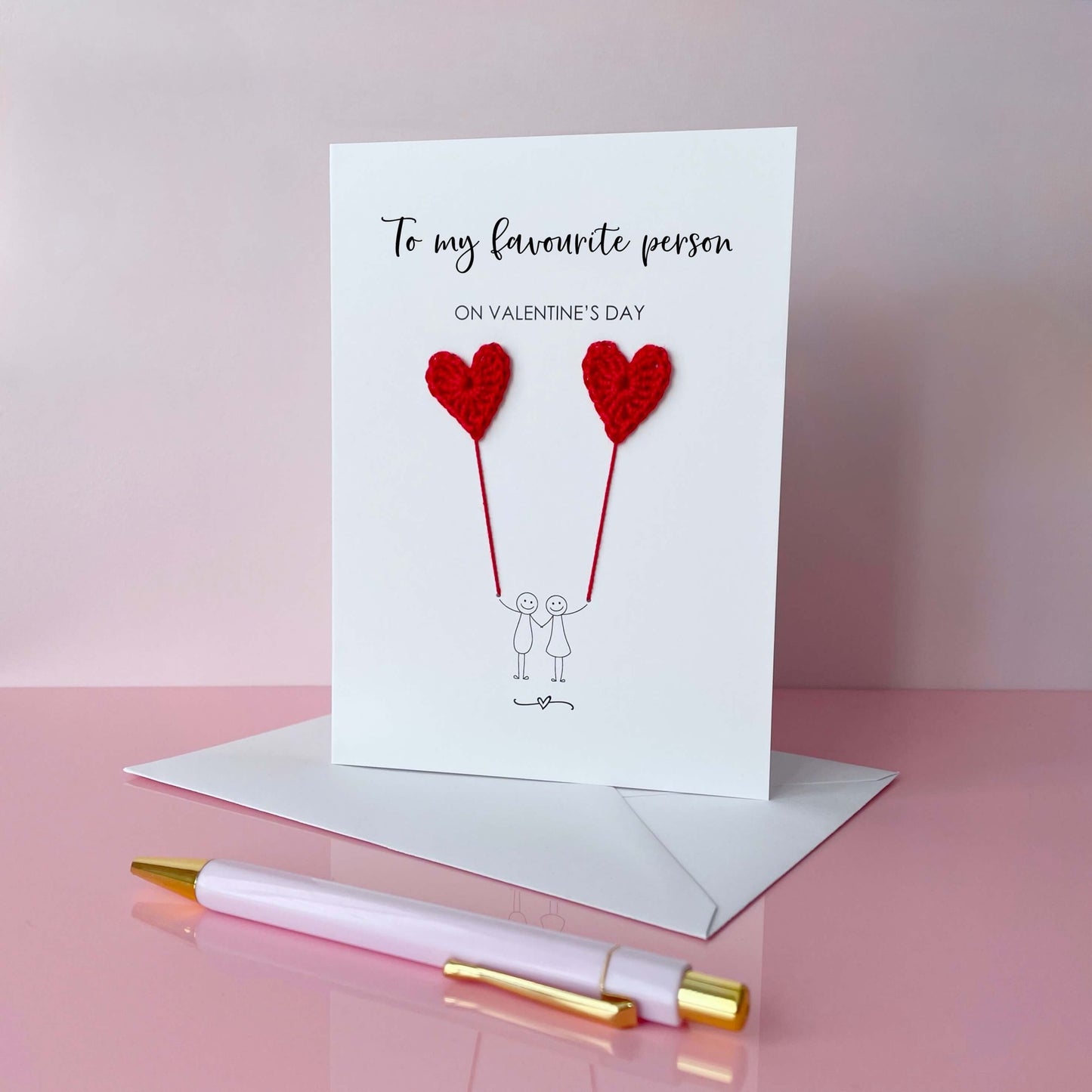 A Valentines card featuring a minimalist design with two figures holding red crochet heart balloons. The text at the top reads: ‘To my favourite person on Valentine’s day’. The card is photographed on a pale pink background.