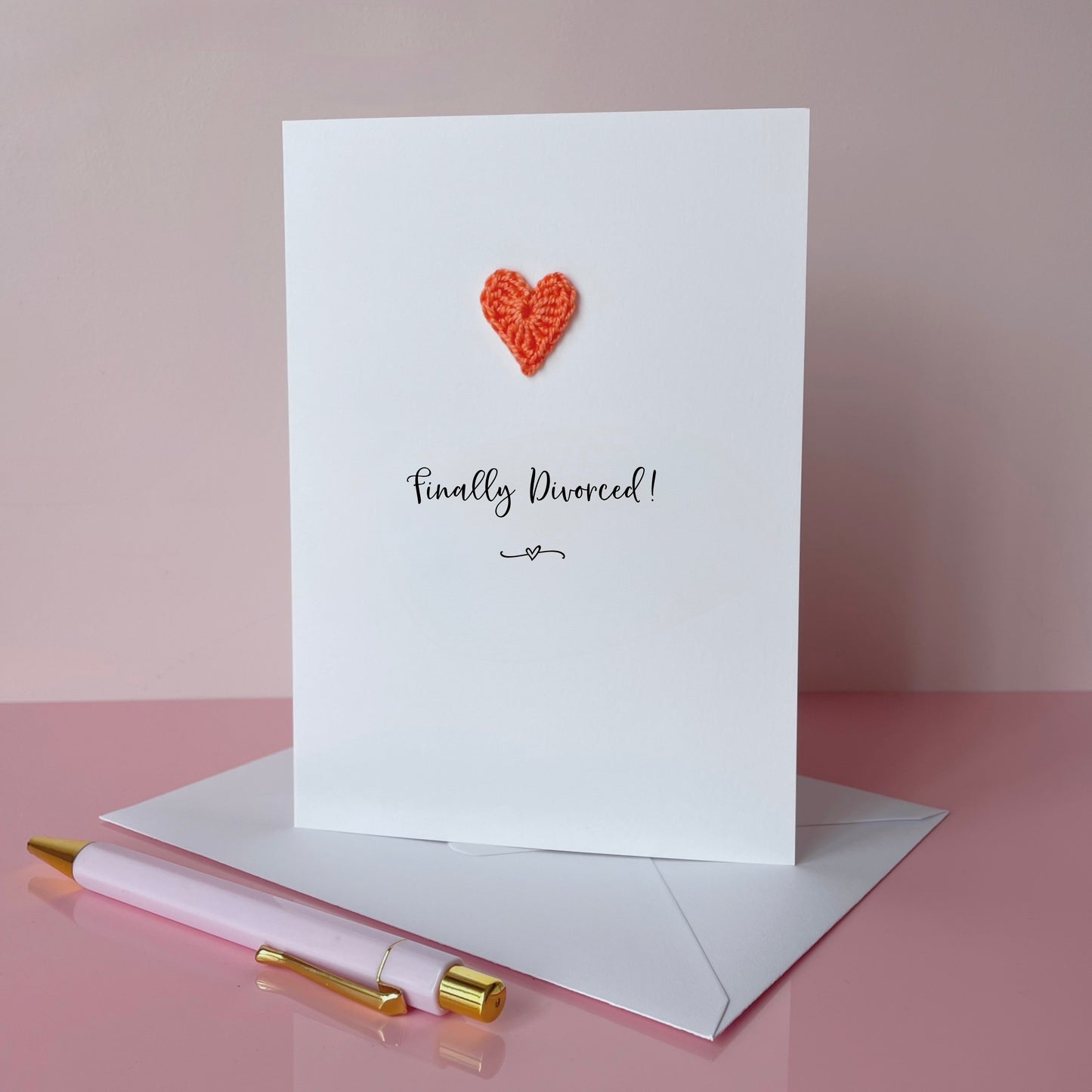 A white card with Finally divorced printed in the centre in black cursive text. A small orange coloured crochet heart has been added to the card above the text. 