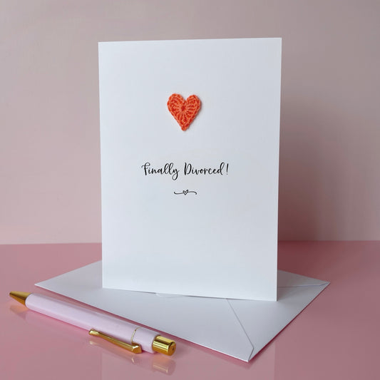 A white card with Finally divorced printed in the centre in black cursive text. A small orange coloured crochet heart has been added to the card above the text. 