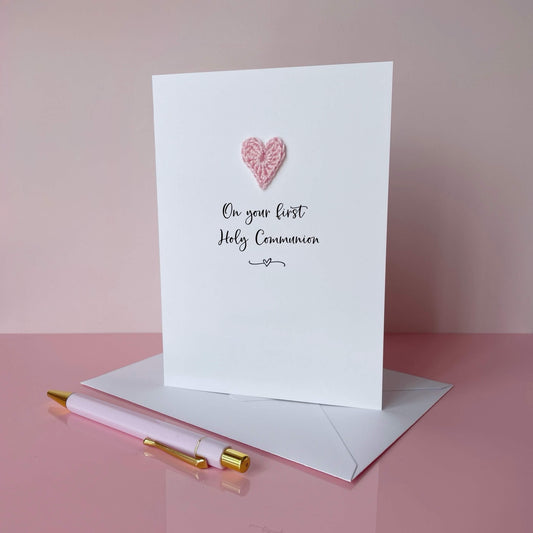 Greeting card with a pale pink crochet heart and text on a pink background, 'On your first Holy Communion' is printed on the card in lovely black cursive text. 