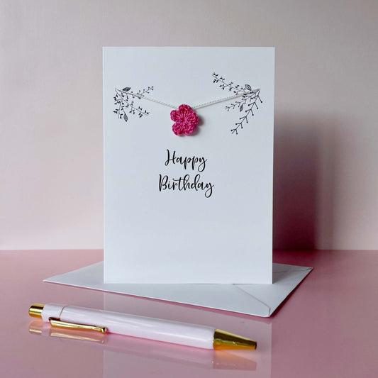 A white card with 'Happy Birthday' printed in the centre of the card in black cursive text. There is some line drawn foliage avove this wording and a bright pink crochet flower has been strung across the card on white yarn.