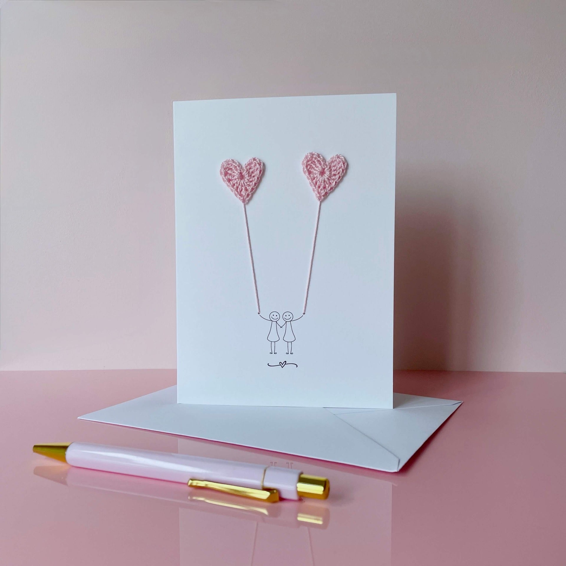 Greeting card with pale pink crochet heart balloons and two women figures on a pink surface with a pen.