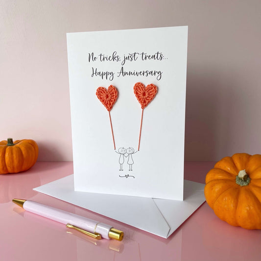 A white card featuring a line drawn couple (complete with pumpkin heads), each holding a orange coloured crochet heart balloon. The wording printed at the top of the card is: No tricks, just treats, Happy Anniversary 
