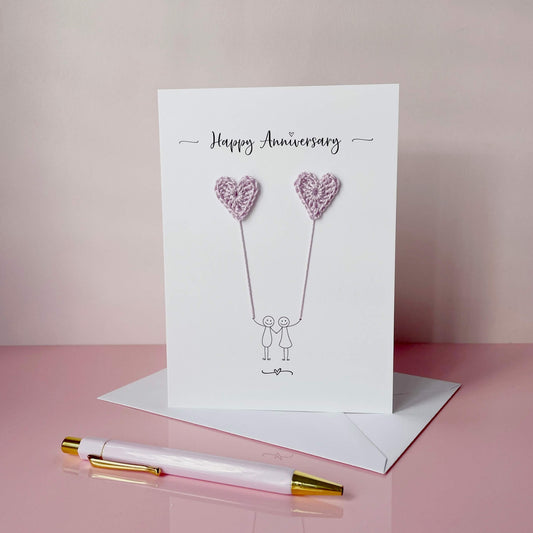 The image shows a beautiful handmade anniversary card with two small lilac coloured crochet hearts, these are attached by yarn, resembling balloons being held by a hand-drawn couple underneath. The text on the card reads "Happy Anniversary" at the top in black cursive text. 